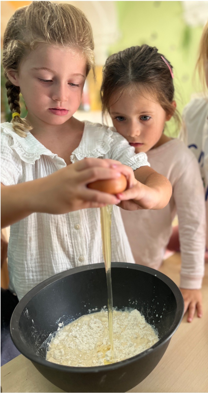 A child cracking an egg into a mixing bowl while a classmate watches